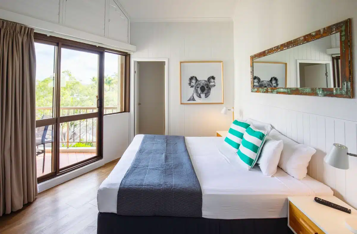 A bright bedroom with a large bed, striped pillows, a dark gray blanket, a wall mirror, a framed koala artwork, and glass doors leading to a balcony with greenery outside.
