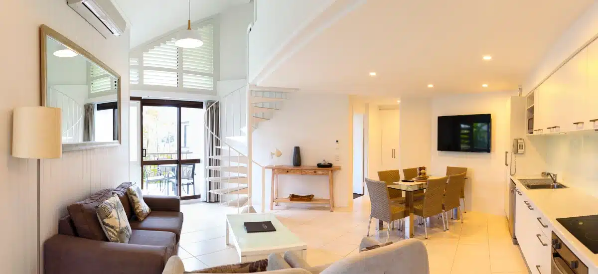 Modern, bright apartment with open-plan living area, spiral staircase, wall-mounted TV, dining table with wicker chairs, kitchen along one wall, large window, and neutral decor.