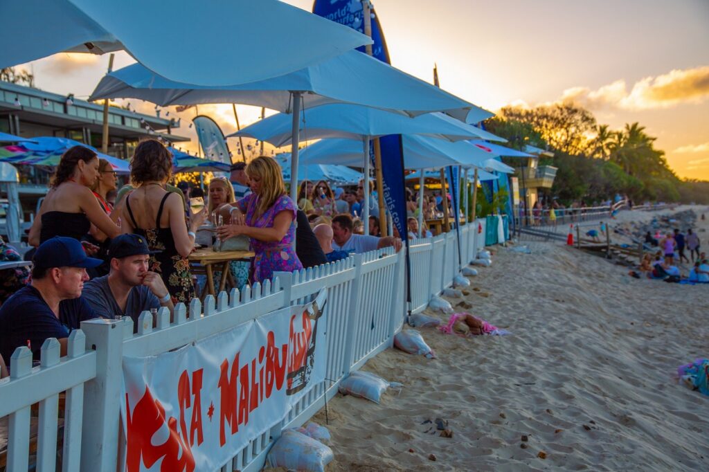 People sit at tables under umbrellas on a patio next to a sandy beach at sunset. Others relax on the sand. A white fence with a "SEA MAIDEN'S" sign separates the patio from the beach.