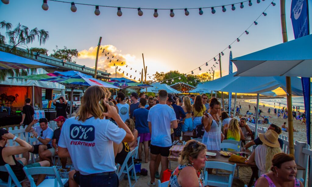 A lively beachside party at sunset with people socializing under string lights and colorful umbrellas, overlooking the ocean. Some are seated at tables, others standing, and the beach is visible in the background.