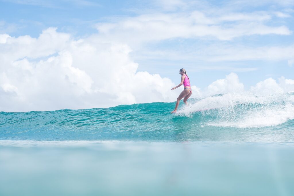 A person wearing a pink swimsuit rides a blue ocean wave on a surfboard under a bright, partly cloudy sky.