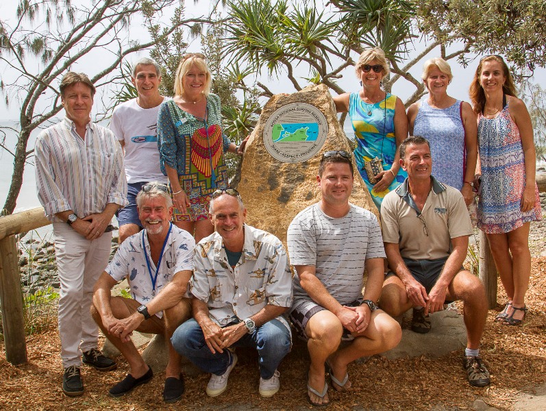 A group of ten adults poses outdoors in front of a large commemorative stone with a colorful plaque, surrounded by trees and plants on a sunny day near the water.