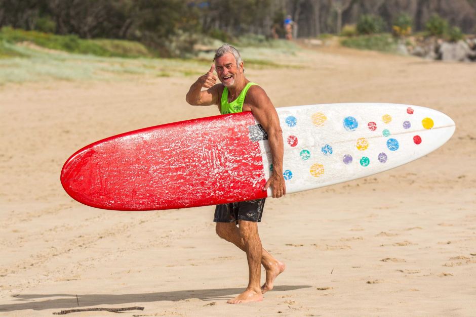 An older man smiles and salutes while carrying a surfboard with a red tip and colorful polka dots, walking barefoot on a sandy beach with greenery in the background.