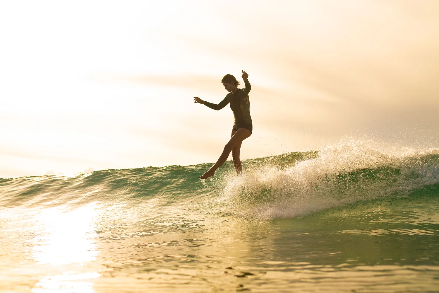 A person in a wetsuit rides a wave on a surfboard at sunset, balancing gracefully with arms outstretched as golden sunlight reflects on the water.