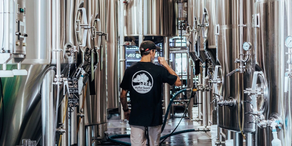 A person wearing a black t-shirt and cap walks between large stainless steel brewing tanks in a brewery, holding a hose and facing away from the camera.