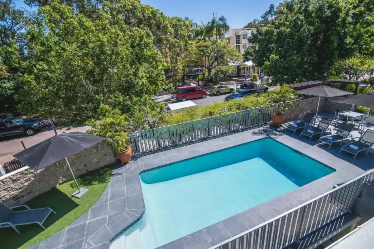A rectangular outdoor swimming pool surrounded by gray tile, lounge chairs, and umbrellas, with a street, trees, and parked cars visible in the background.