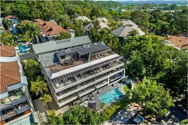 Aerial view of a modern multi-story building with balconies and a pool surrounded by lush trees and neighboring houses with red and gray roofs. The setting appears tropical and green.