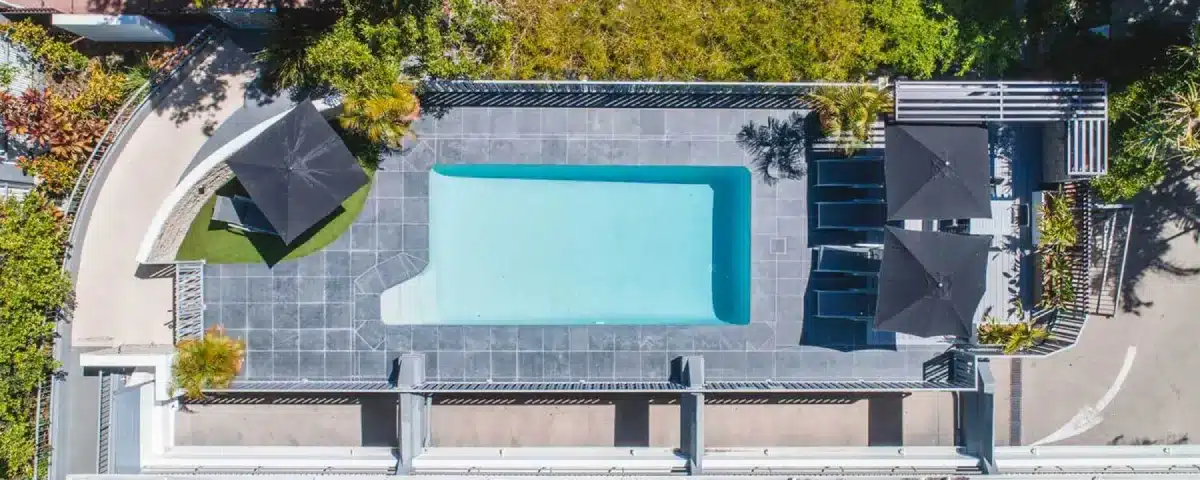 A rectangular outdoor swimming pool with clear blue water, surrounded by gray tiles, black lounge chairs, and black umbrellas, viewed from above. Green plants and landscaping line the pool area.