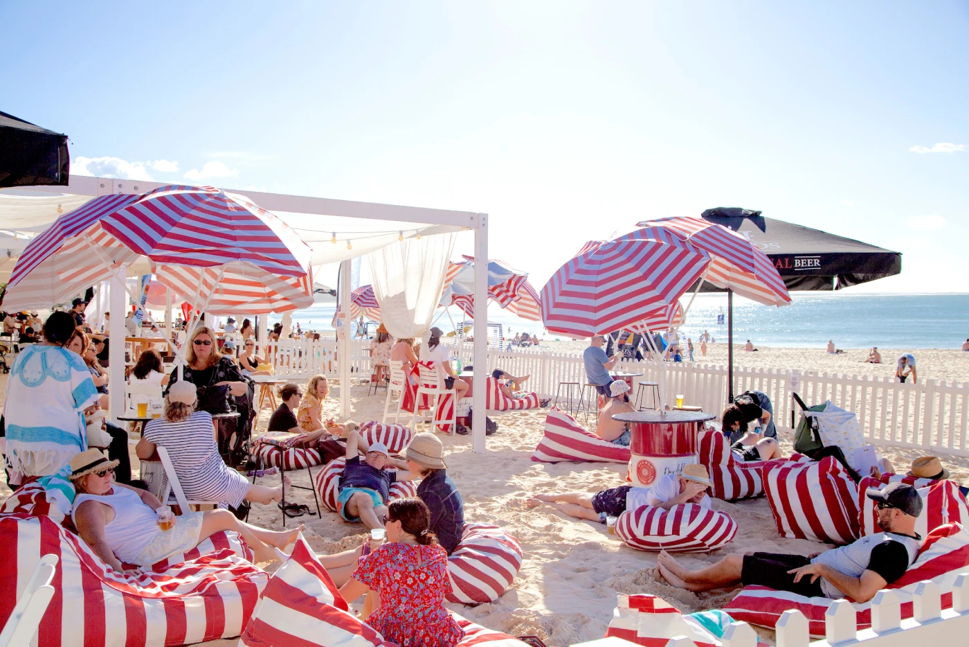 People relaxing on a sunny beach under red and white striped umbrellas, lounging on bean bags and chairs; a white fence, the ocean, and a bright sky are in the background.