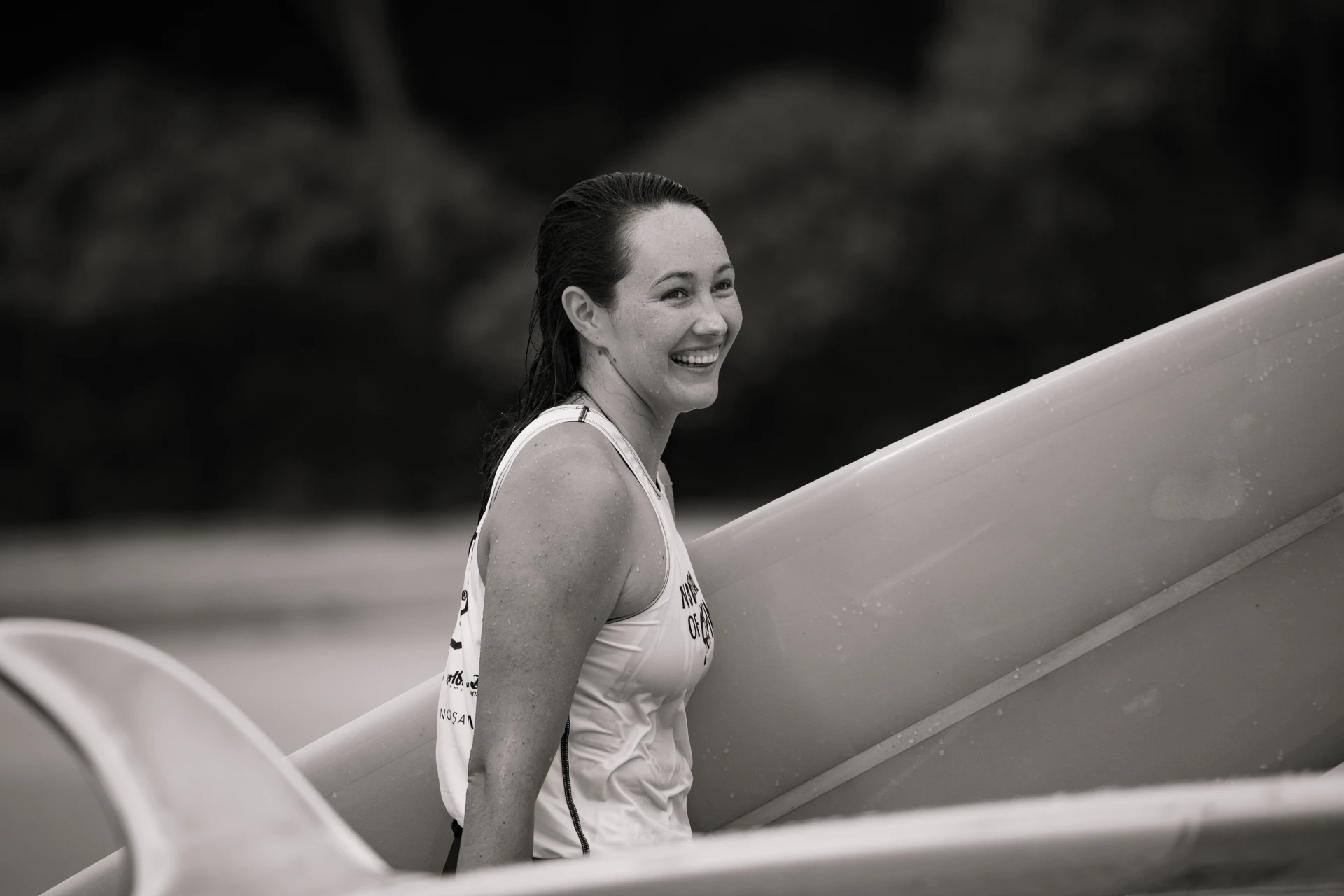 A woman with wet hair smiles while carrying a surfboard on the beach. She wears a sleeveless top and appears happy, with blurred trees in the background. The photo is in black and white.