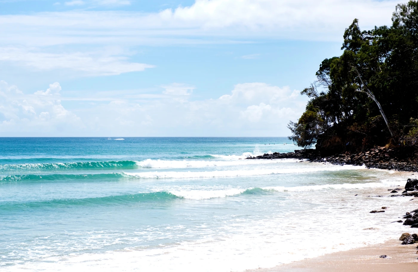 A sunny beach scene with gentle turquoise waves rolling onto sandy shore. To the right, green trees and rocky outcrops extend into the water under a partly cloudy blue sky.