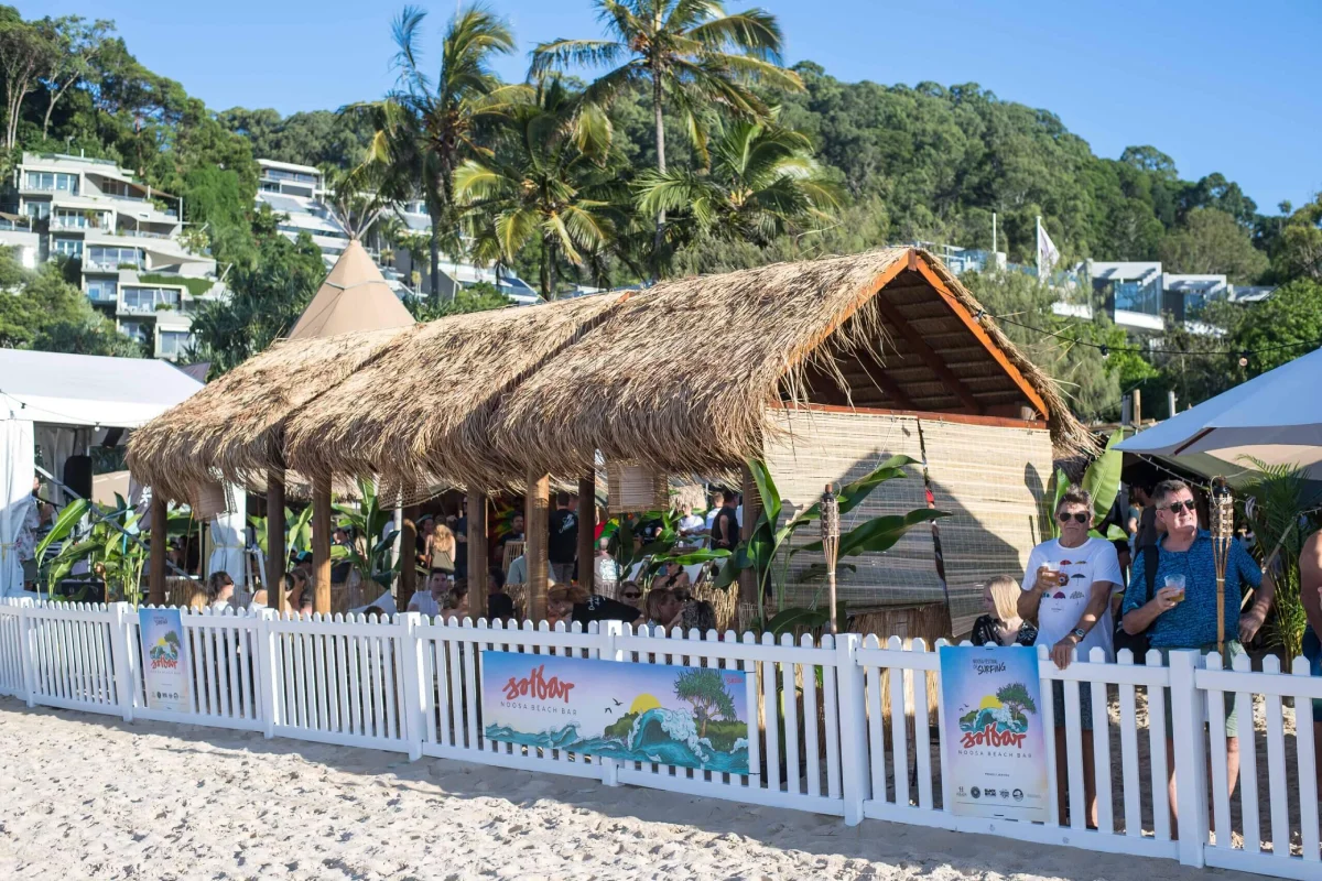 A thatched-roof beach hut surrounded by tropical plants sits behind a white picket fence, with people gathered outside. Modern buildings and palm trees are visible in the background under a clear sky.