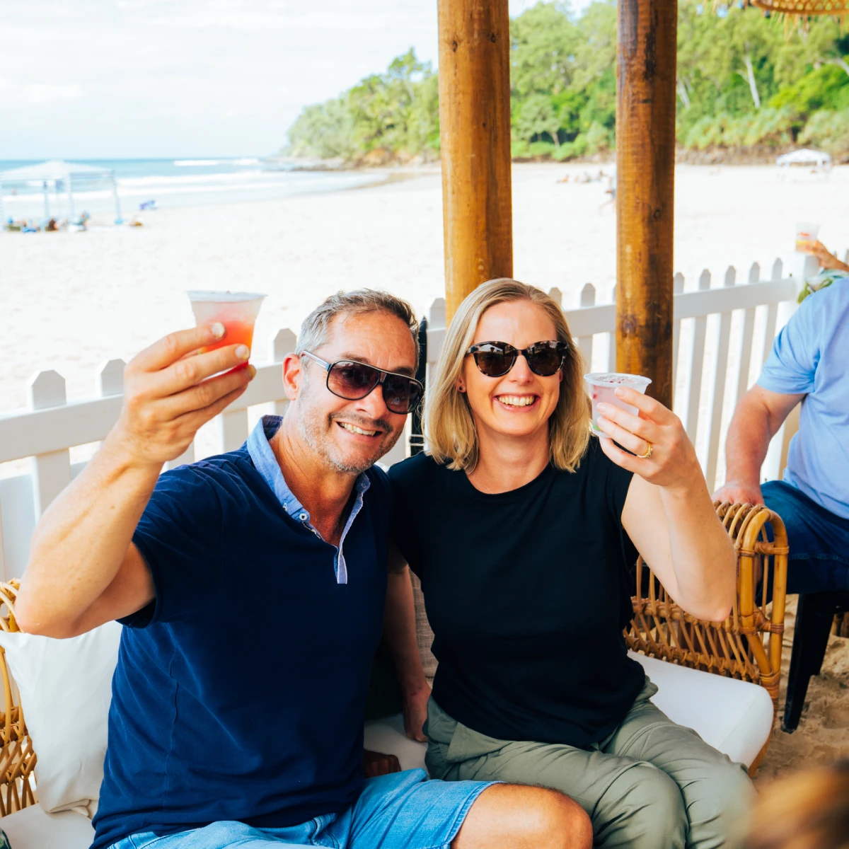 A smiling man and woman wearing sunglasses sit on a beachside patio, holding up drinks and posing for a cheerful photo. The sandy beach and ocean are visible in the background.