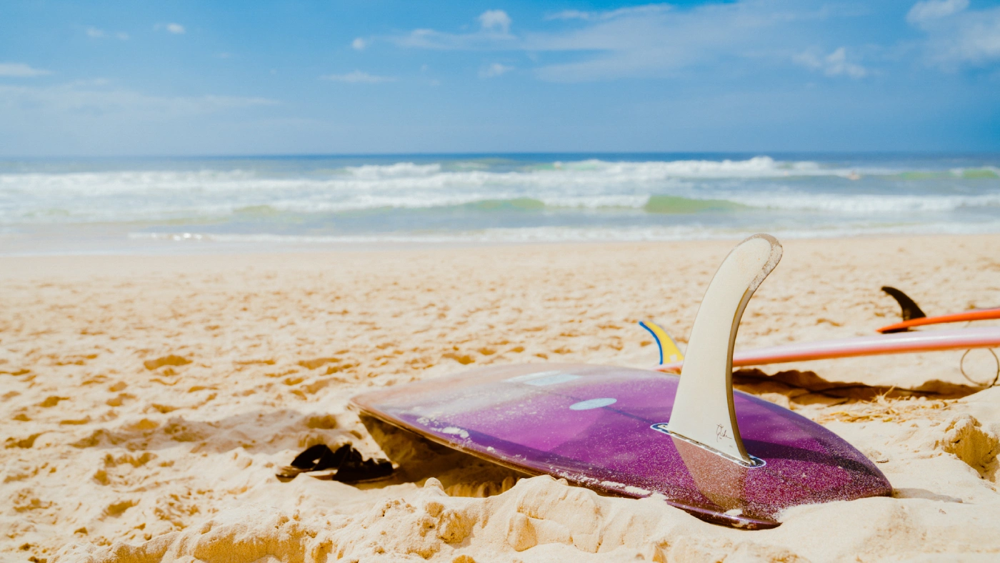 A purple surfboard lying fin up on sandy beach near the shoreline, with ocean waves and blue sky in the background; another surfboard is partially visible to the right.