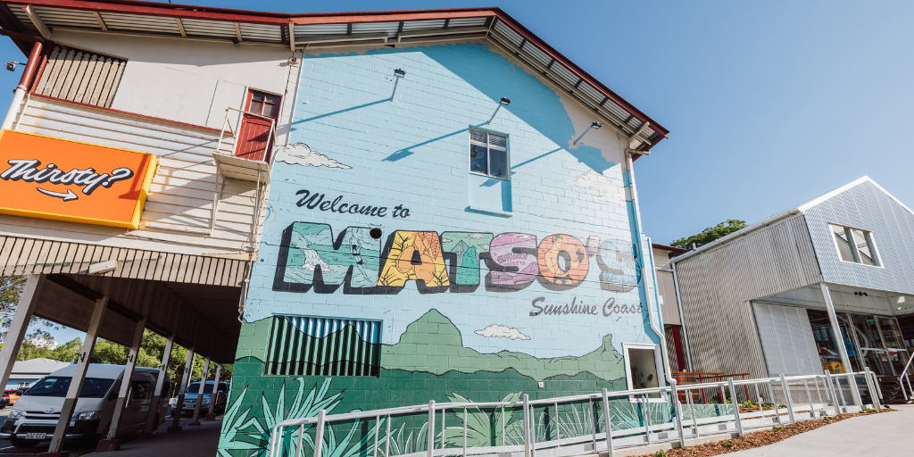 A large colorful mural on a building wall reads "Welcome to MATSO'S Sunshine Coast" with a scenic background of hills, clouds, and greenery. A bright orange "Thirty?" sign is visible on the left.