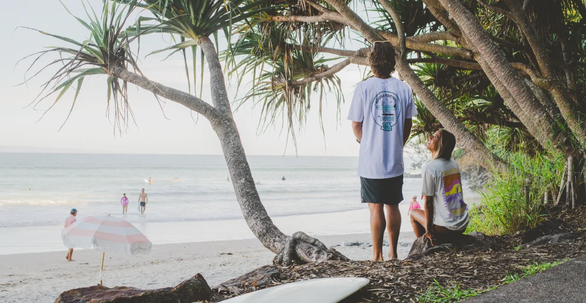 Two people under trees by a beach, one standing and one sitting on rocks, watching the ocean. A surfboard lies on the ground nearby. In the distance, people walk and play near the water under a striped beach umbrella.