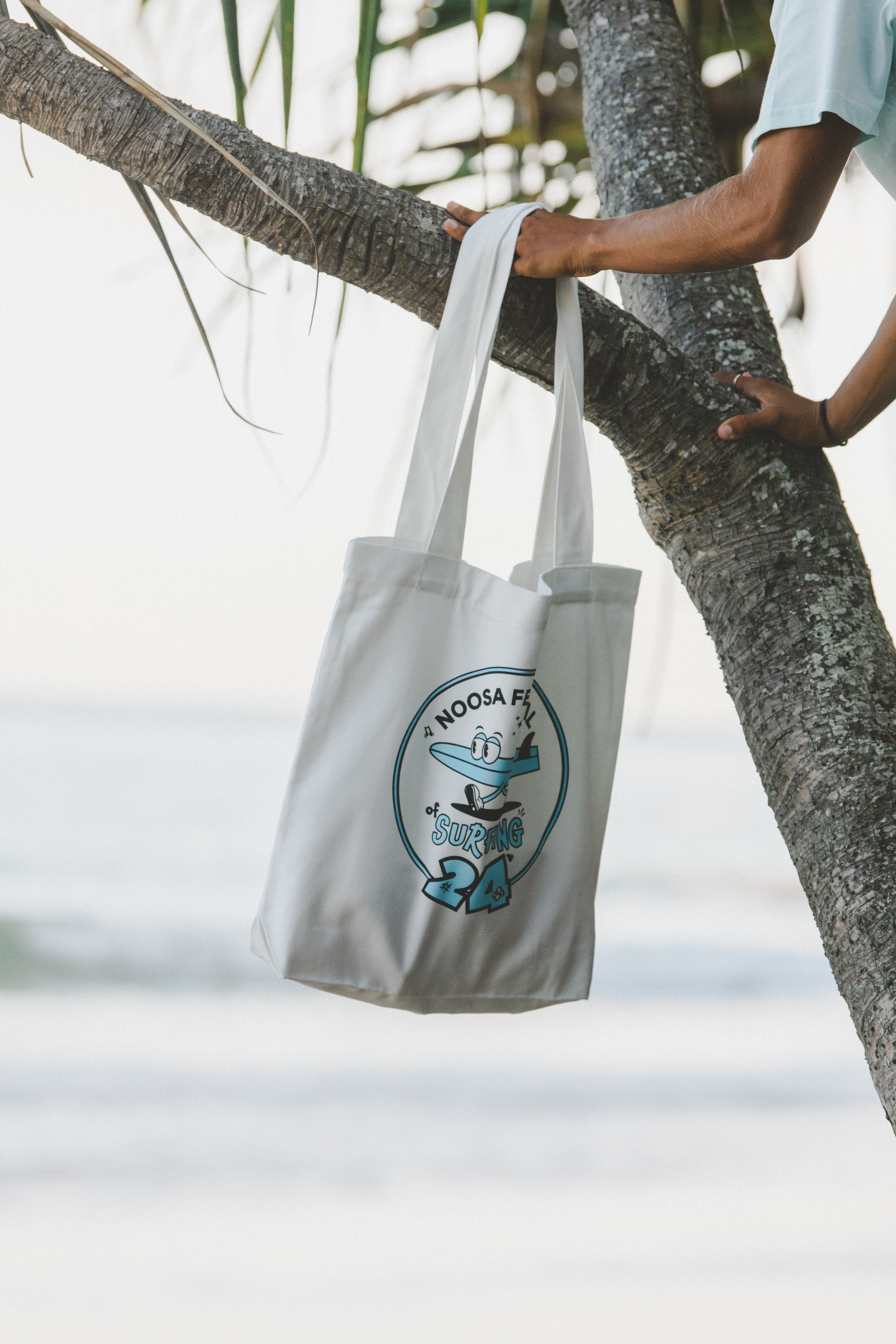 A person hangs a white tote bag with a surfing cartoon graphic and the text "Noosa Festival of Surfing 24" on a tree branch near a beach.