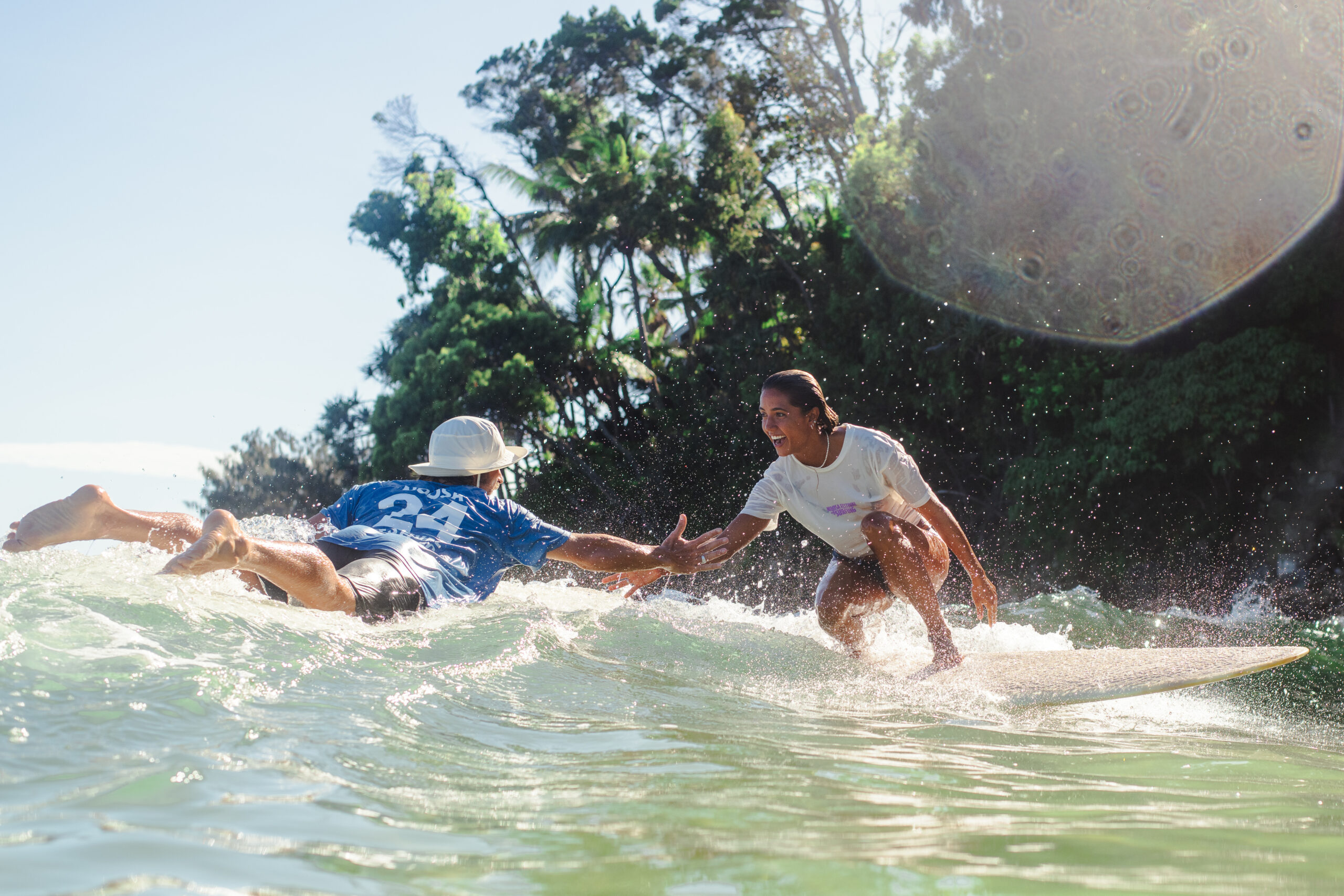 Two surfers, one paddling and one riding a wave, reach out for a high-five in the ocean near lush green trees and bright sunlight.