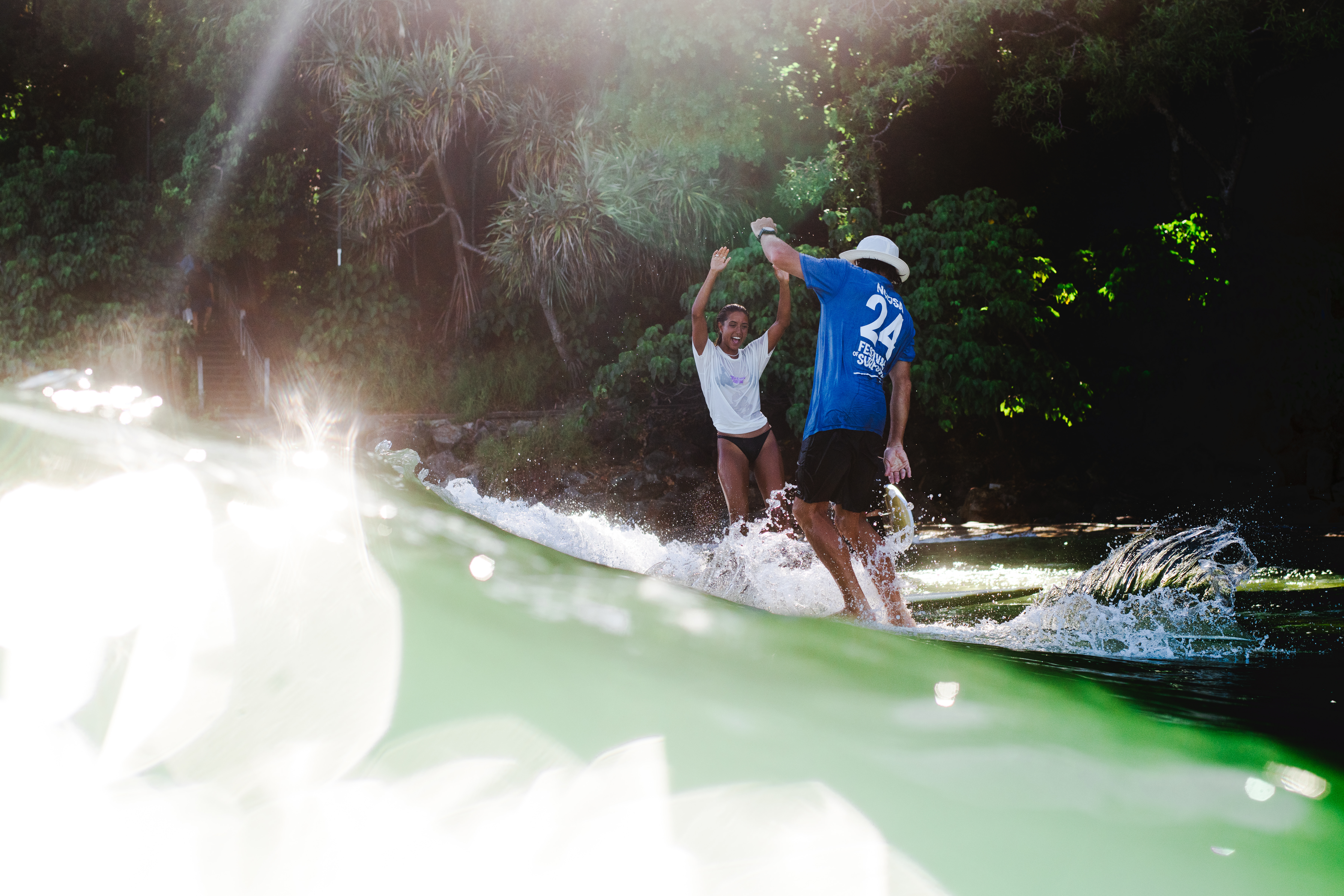 Two people riding a wave on surfboards give each other a high five near the shore, surrounded by lush greenery and sunlight reflecting off the water.
