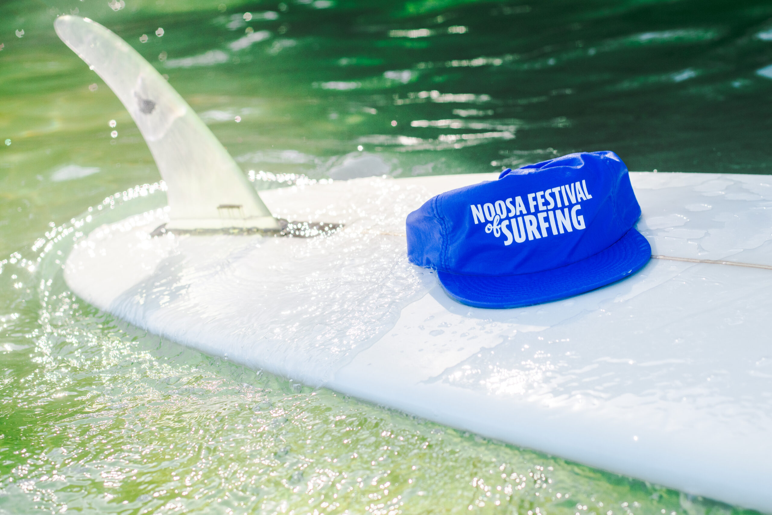 A blue hat with "Noosa Festival of Surfing" printed on it rests on a white surfboard floating in greenish water, with the surfboard’s fin visible in the sunlight.
