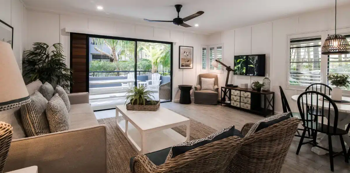 A stylish living room with wicker chairs, a gray sofa, plants, a white coffee table, and a TV. Large glass doors open to a patio, and natural light fills the room. A ceiling fan hangs above and a dining table sits by the window.