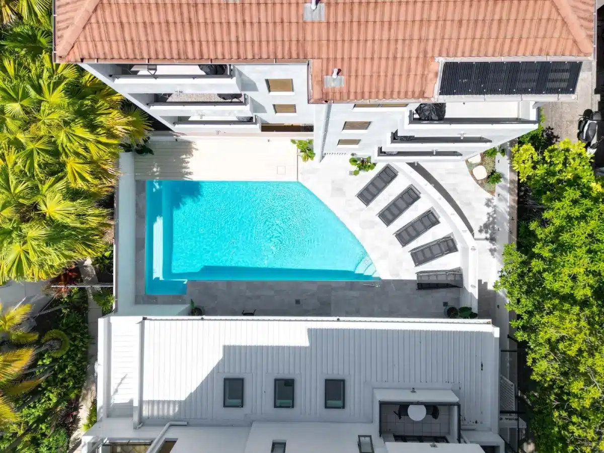 Aerial view of a rectangular outdoor swimming pool with clear blue water, surrounded by lounge chairs on a white deck, flanked by buildings and greenery.