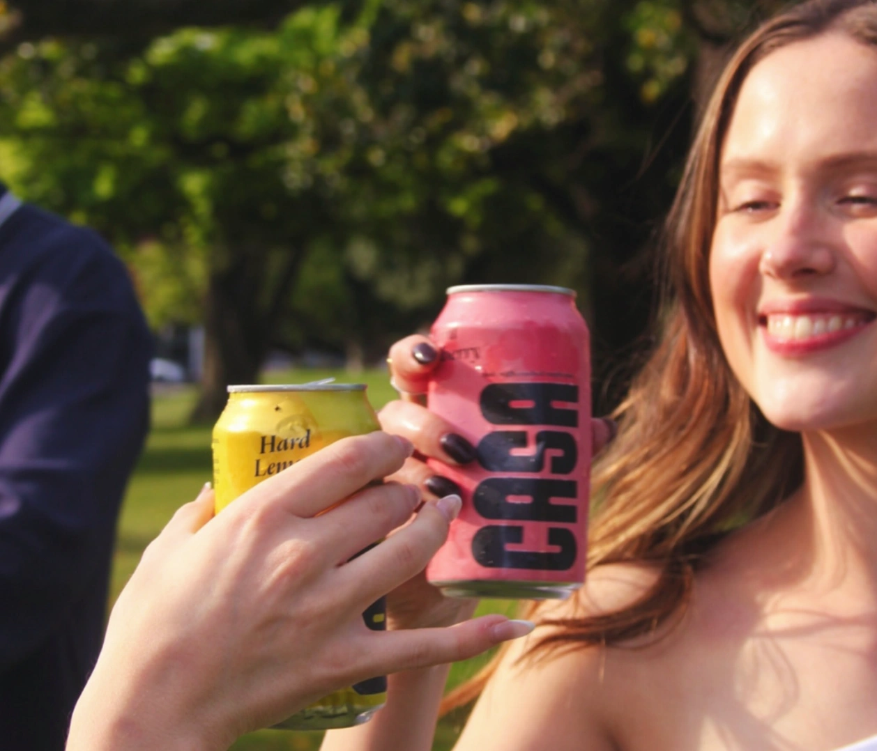 Two people clink cans outdoors; one holds a pink CASA can, the other a yellow hard lemonade. A woman smiles in the sunlight with trees in the background, suggesting a cheerful, casual outdoor gathering.