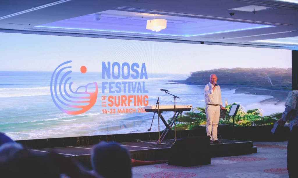A man stands on stage with a microphone in front of a large screen displaying “Noosa Festival of Surfing 14-23 March 2019” and an ocean scene, with musical equipment beside him.