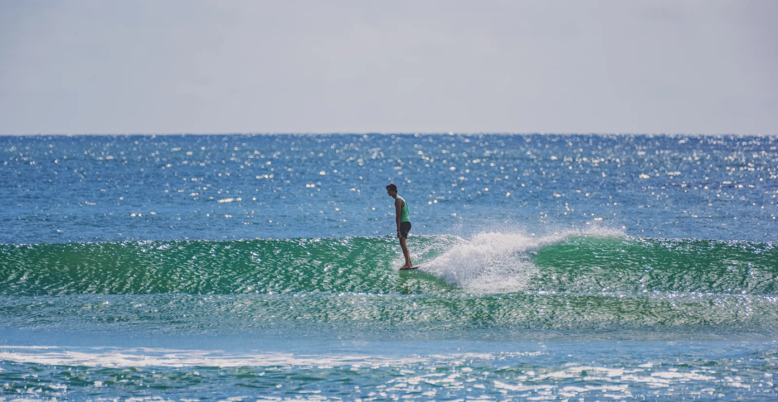 A person in a green shirt is surfing on a small, clear wave in the ocean under a bright, sunny sky. The water is sparkling in the sunlight.