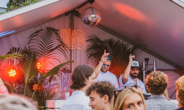 Two DJs perform under a white tent decorated with plants and a disco ball, with vibrant lights and a neon sign, as a crowd of people enjoy the outdoor event.