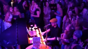 A musician in a hat plays a keyboard at a lively, crowded indoor venue surrounded by people standing close, some taking photos and videos under purple and pink stage lighting.
