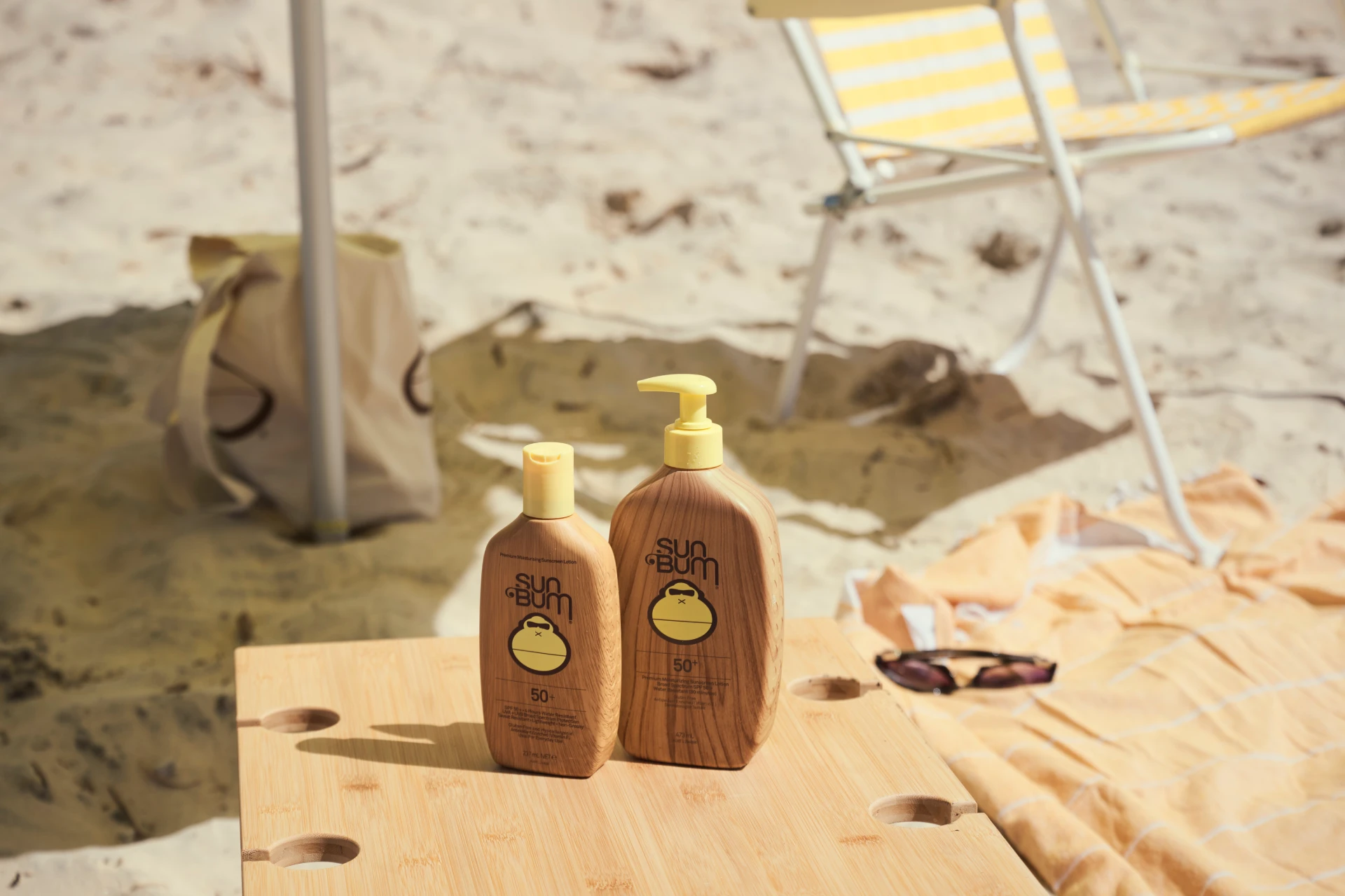 Two bottles of Sun Bum sunscreen sit on a wooden table at the beach, with sand, a striped yellow and white beach chair, a towel, sunglasses, and a beach bag in the background.