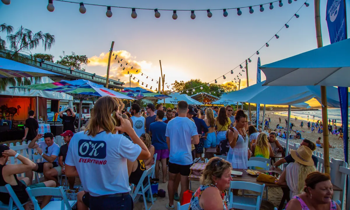 A lively beachside gathering at sunset, with people socializing under string lights and umbrellas near the sand. Some are seated, others stand and chat. The sky is colorful, and the atmosphere is festive and relaxed.