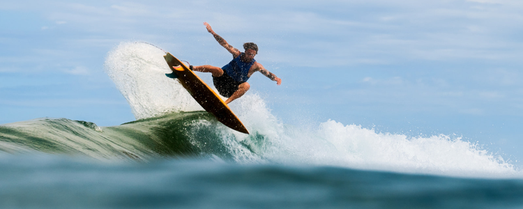 A surfer in a blue shirt catches air above a wave, balancing on a black surfboard with water spraying behind him under a partly cloudy sky.