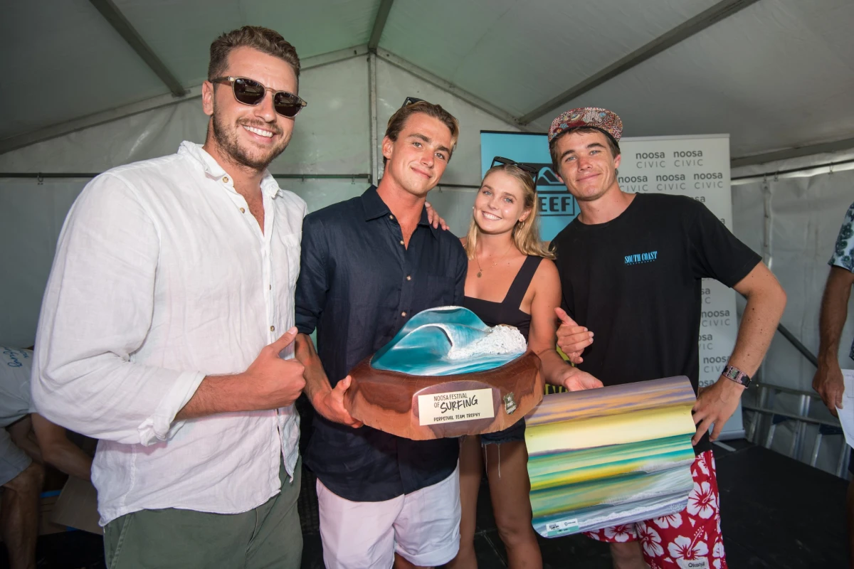 Four young adults stand together under a tent, smiling at the camera. One holds a large wave-shaped trophy, while another holds a colorful rolled-up item. Event signage is visible in the background.