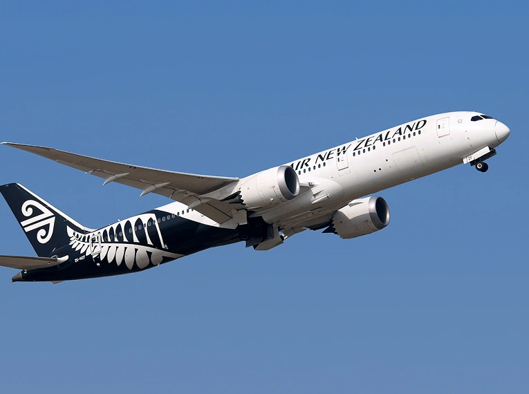An Air New Zealand airplane with a black and white fern design on its tail takes off against a clear blue sky.