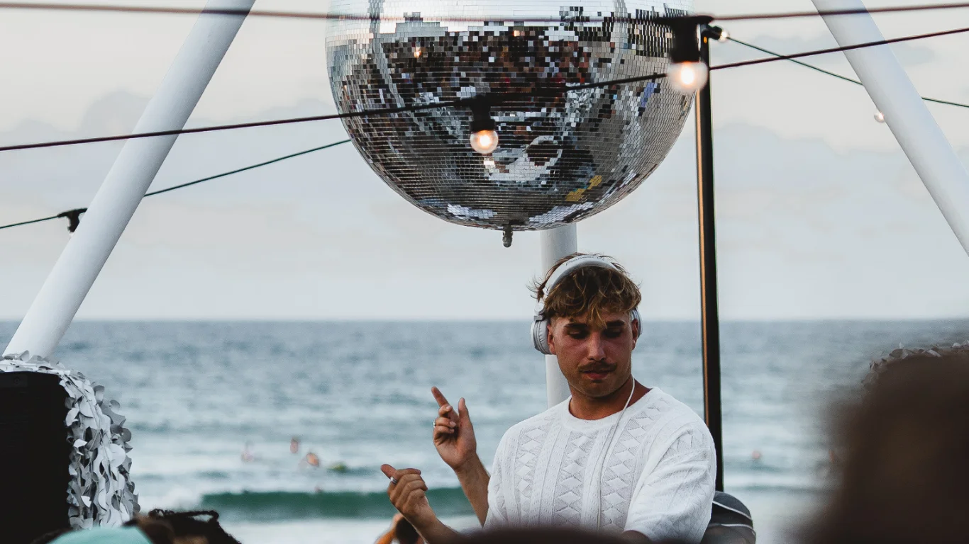 A DJ wearing headphones performs outdoors in front of the ocean, with a large disco ball and hanging string lights above him, while people watch from the foreground.