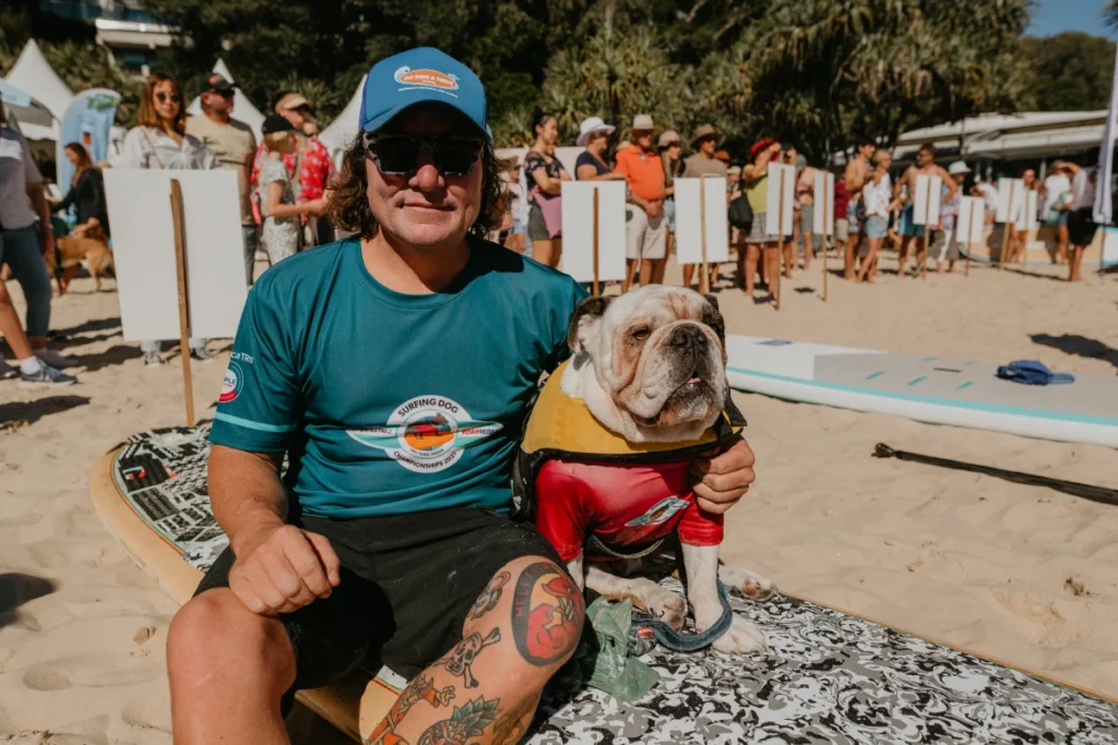 A man in a blue shirt and cap sits on a surfboard in the sand next to a bulldog wearing a yellow and red vest. There is a crowd of people and surfboards in the background.
