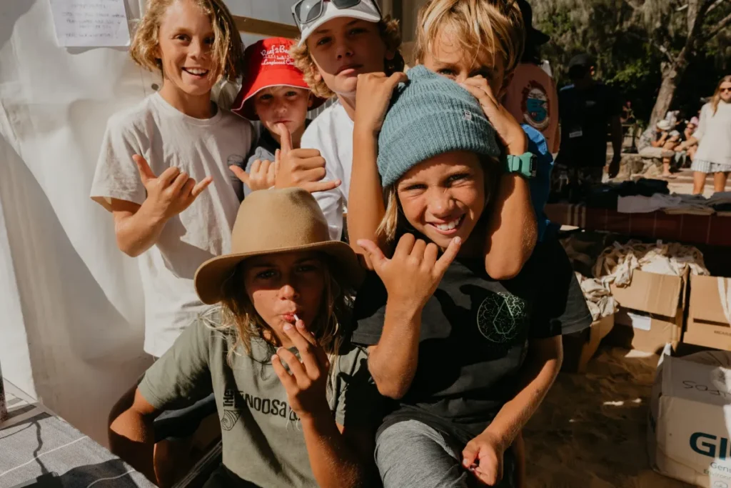 A group of smiling children pose outdoors, making shaka hand signs. They wear casual clothes and hats, with sunlight highlighting their faces. Some lean in close together, creating a fun, lively atmosphere.