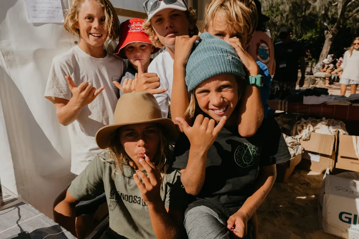 A group of smiling children pose outdoors, making shaka hand signs. They wear casual clothes and hats, with sunlight highlighting their faces. Some lean in close together, creating a fun, lively atmosphere.