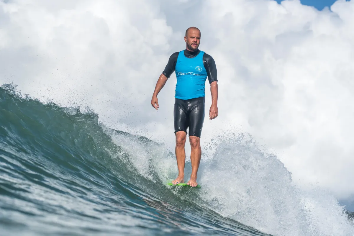 A man in a blue wetsuit top and black shorts is surfing on a wave, standing upright on a green surfboard with clouds and blue sky in the background.