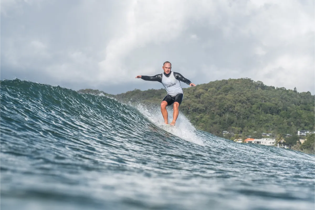 A person in a wetsuit rides a wave on a surfboard near a lush, green shoreline under a cloudy sky, with buildings visible in the distance.