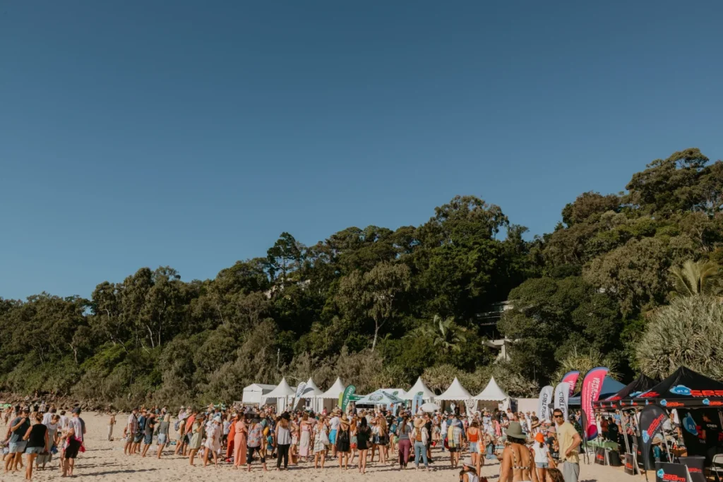 A large crowd gathers on a sandy beach near rows of white tents and colorful banners, with a forested hillside and clear blue sky in the background.