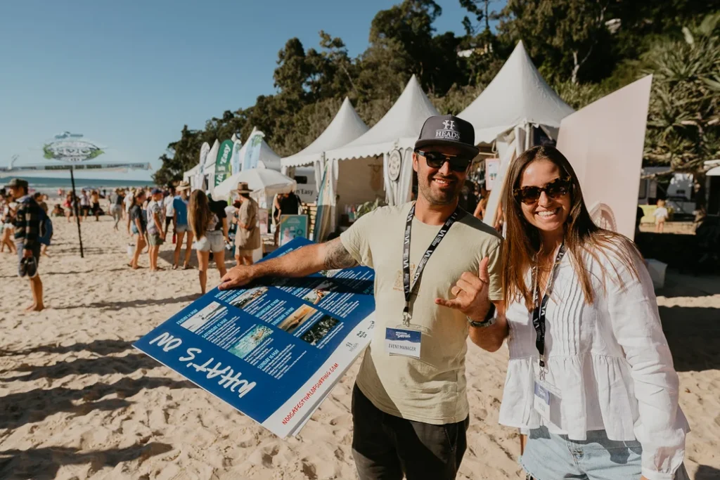 Two people smiling at a beach event, one holding a large information board. Tents and other people are visible in the background on the sandy beach, with trees and blue sky.