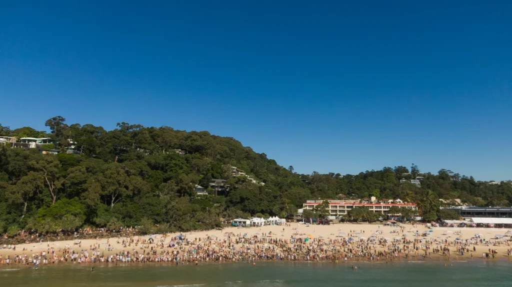 A crowded beach with many people on the sand and near the water, surrounded by lush green trees and buildings under a clear blue sky.