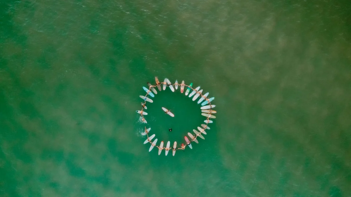 Aerial view of surfers forming a circle with their boards on calm green ocean water, creating a united ring shape with one person in the center.