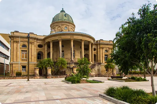 Historic neoclassical building with a large central dome, tall columns, and arched windows, surrounded by trees and greenery in an open plaza under a partly cloudy sky.
