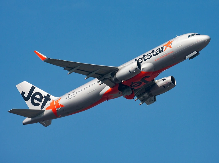 A Jetstar airplane with orange and gray livery ascends against a clear blue sky, its landing gear retracted and engines visible under the wings.