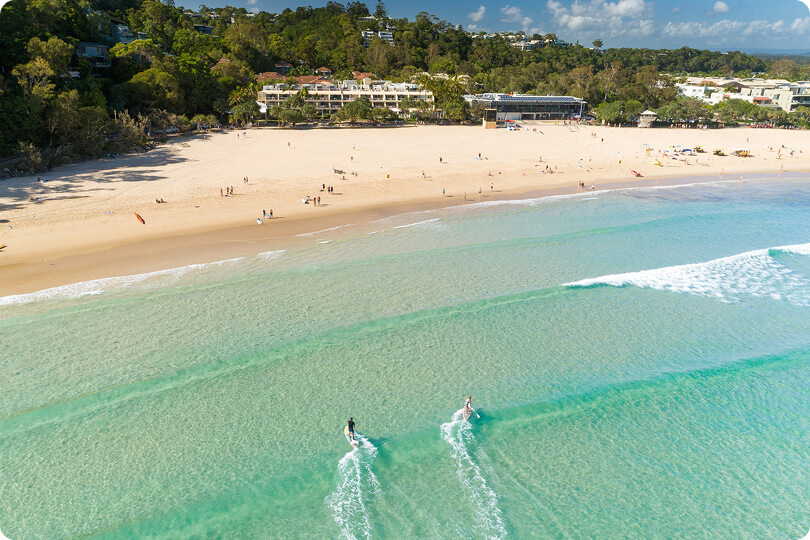 Aerial view of a sandy beach with clear turquoise water, two people paddleboarding near shore, and several people scattered on the sand; buildings and trees in the background.