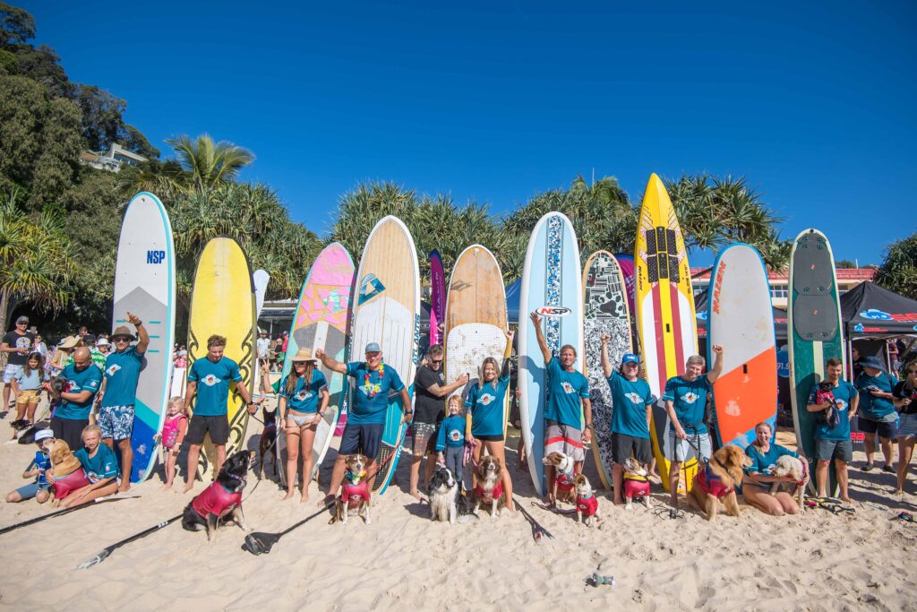 A group of people and dogs pose with colorful surfboards on a sandy beach under a clear blue sky, surrounded by palm trees and beachgoers.